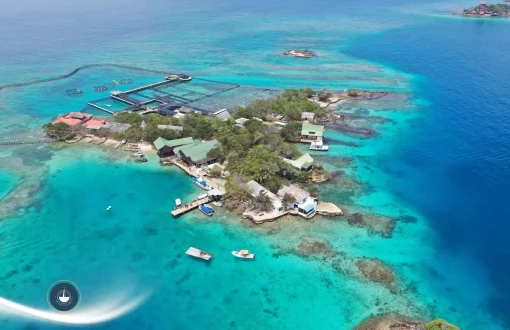 Vista aérea de una pequeña isla tropical rodeada por aguas cristalinas de tonos turquesa y azul profundo. La isla cuenta con construcciones costeras, muelles, embarcaciones y áreas marinas delimitadas, en medio de un entorno coralino en el Caribe Islas del Rosario en yate: así se vive el archipiélago de verdad navega colombia